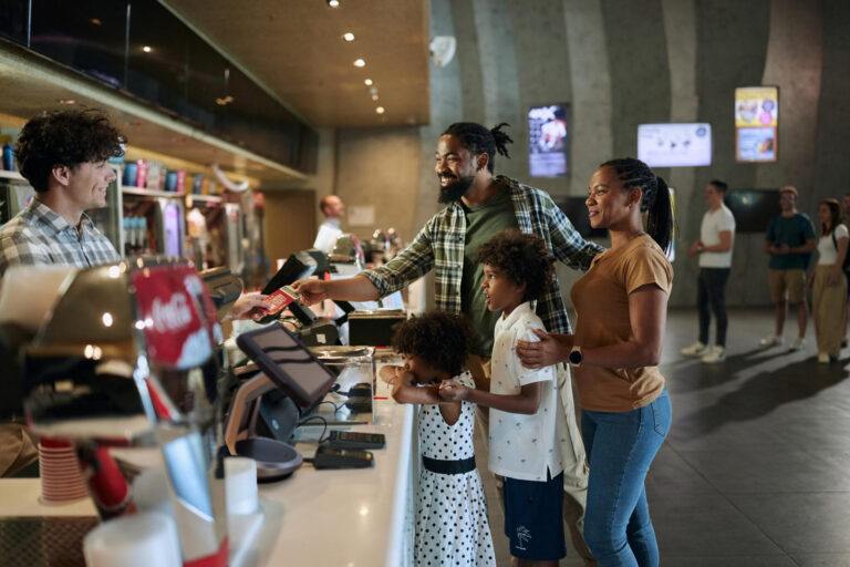 Felices padres afroamericanos y sus hijos comprando entradas en un puesto de comida en el estadio de artículos deportivos.